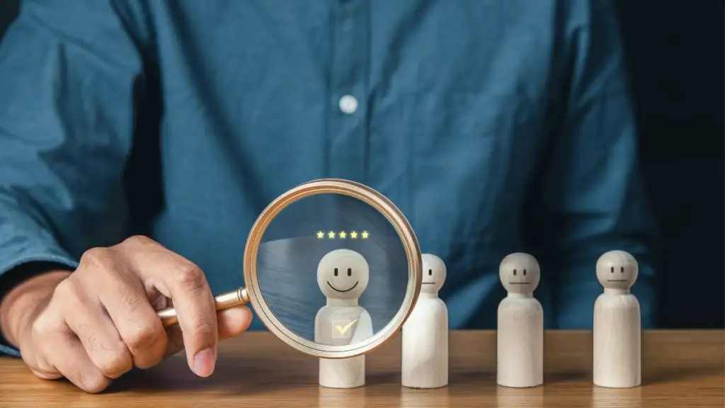 AI in HR - Person examining wooden figures with magnifying glass, highlighting a smiling figure with five stars above it